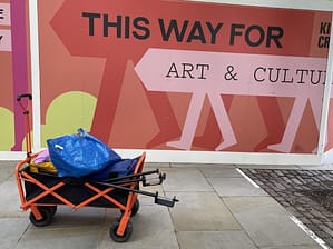 A camping wagon full of bags. Behind is a bright hoarding with the text. 'This Way For Art & Culture'