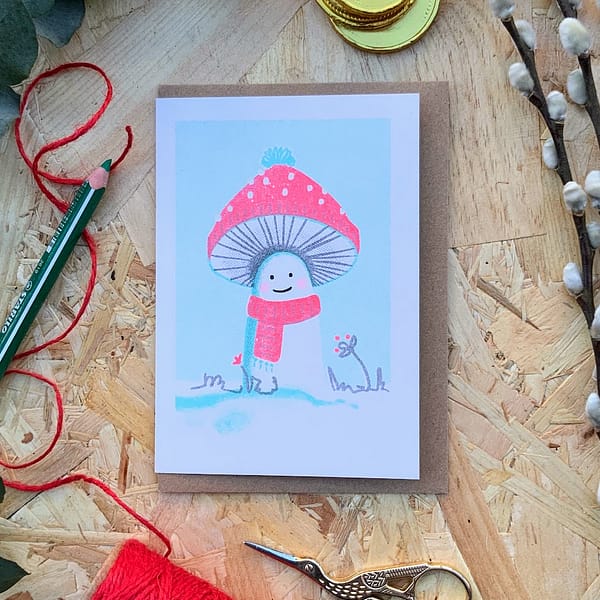 A photograph of a risograph print christmas card with a 'Cosy Mushroom' character wearing a woolly hat and scarf - ready for some fun in the snow. 